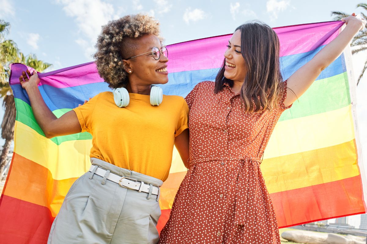 Two people smiling and holding up a rainbow pride flag behind them.