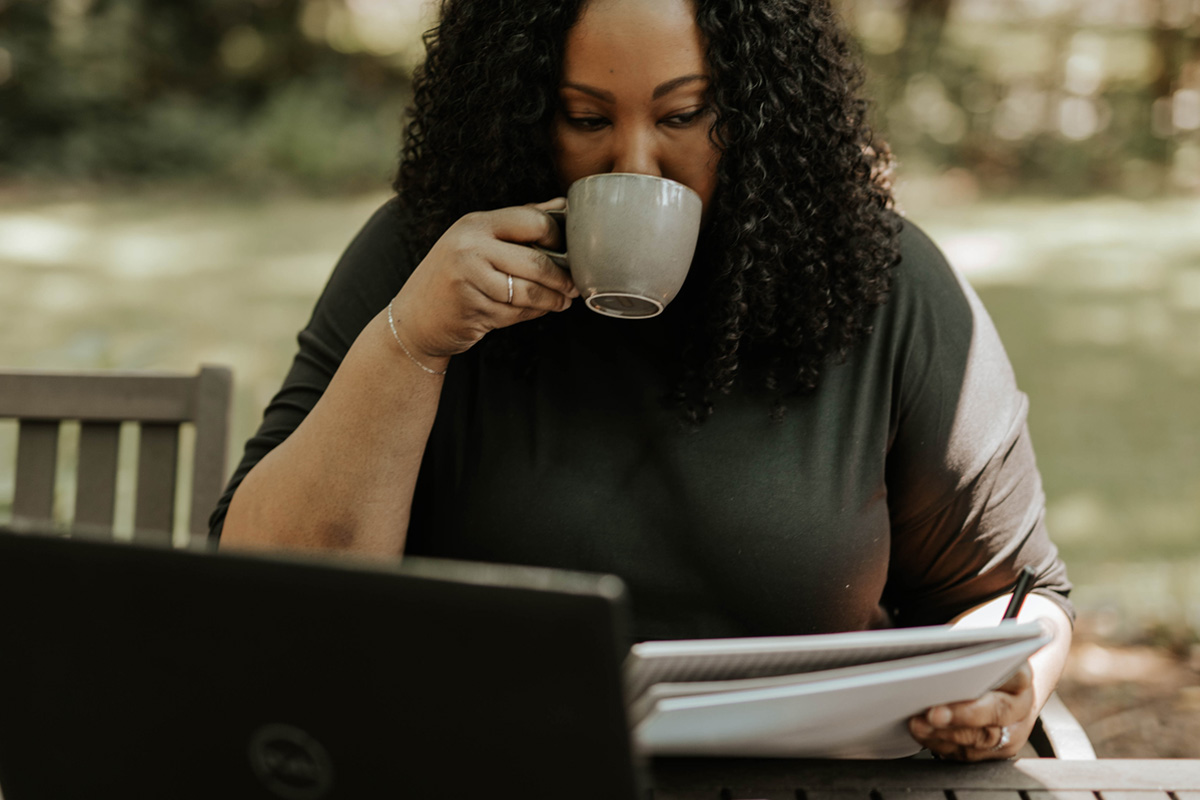 A Black woman sitting outside, sipping something out of a mug, with on her computer and holding a notepad.