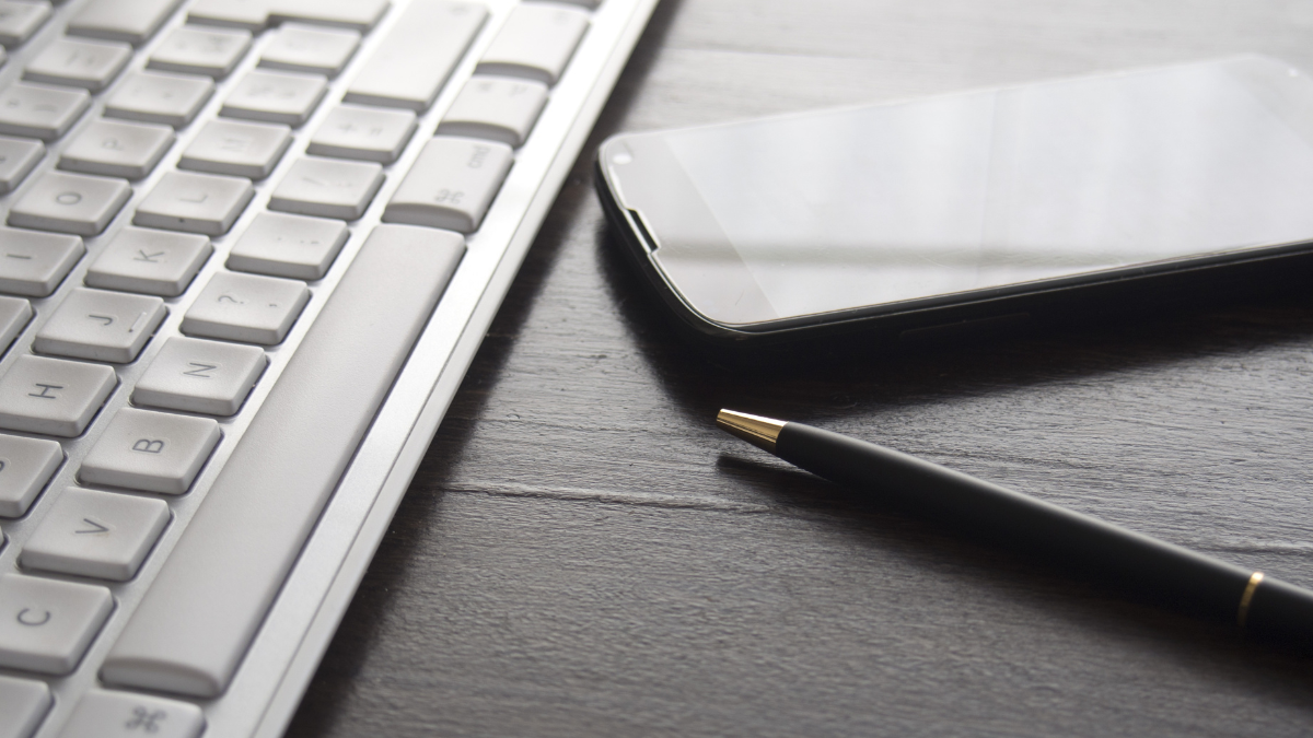 Monochrome photo of a keyboard, a smart phone, and a pen on a desk.