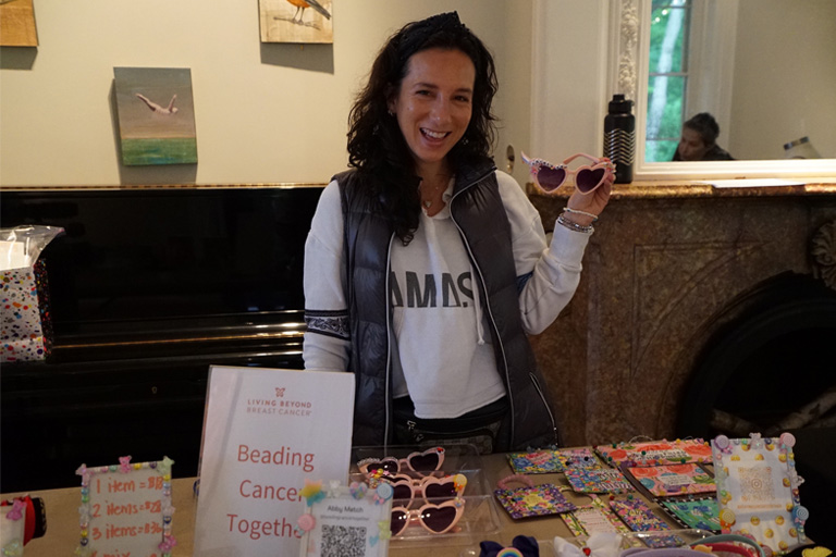 Woman smiling, holding heart-shaped sunglasses, at her fundraiser table. The sign reads, "Beading Cancer Together."