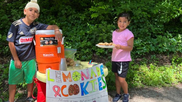 Two children smiling next to their lemonade stand, one is holding a plate of cookies. Their handmade sign reads, "Lemonade & cookies."