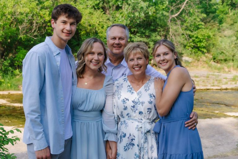 Tammy Uhl is surrounded by her husband and their three grown children, all wearing shades of blue and white in a tranquil outdoor setting