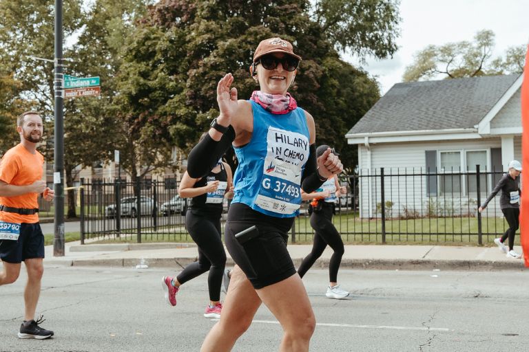 Hilary Weckstein waves as she runs in a marathon in an urban setting.